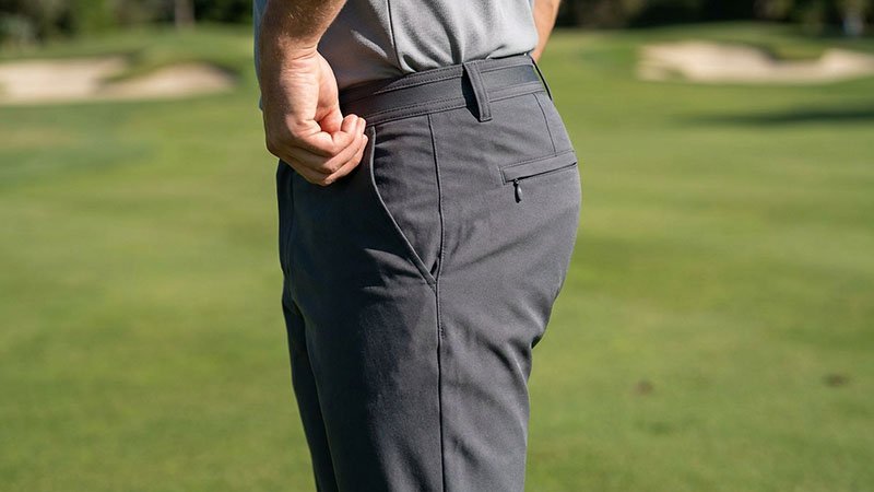 A golfer reaching into the pocket of well-fitting golf pants, showing the stretch fabric and a secure back pocket